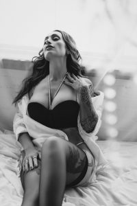 Brunette woman with tattoos in black lingerie and white button up shirt laying on bed and leaning against window