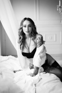 Beautiful brunette woman with tattoos, in black lingerie and white button up top, on a bed with white sheets, white textured wall.